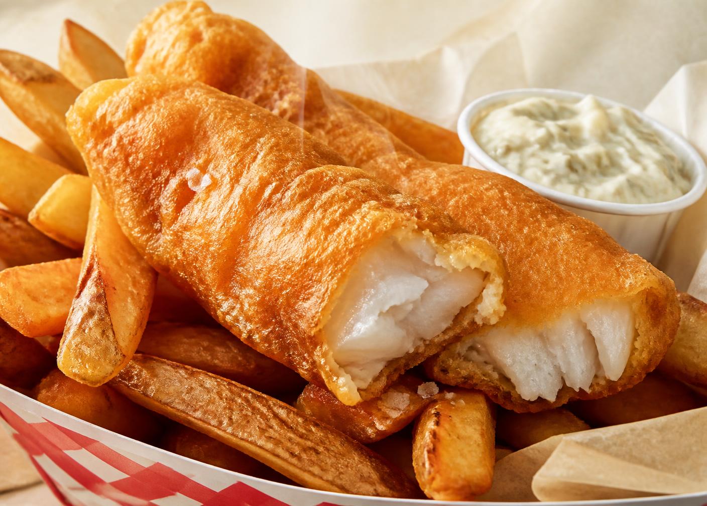 Hand-battered fish fillet broken open to show flaky white pollock, beside thick hand-cut fries and tartar sauce